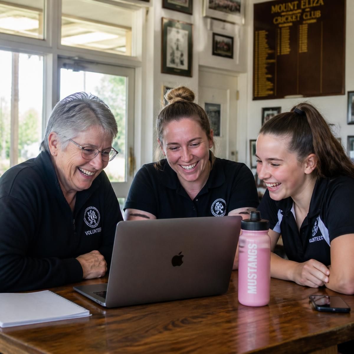 Three club volunteers of different ages laughing together around a laptop in a clubhouse